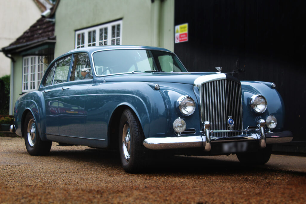 Powder Blue Bentley S1 Continental flying spur parked on a beige gravel driveway in from of a pastel green building