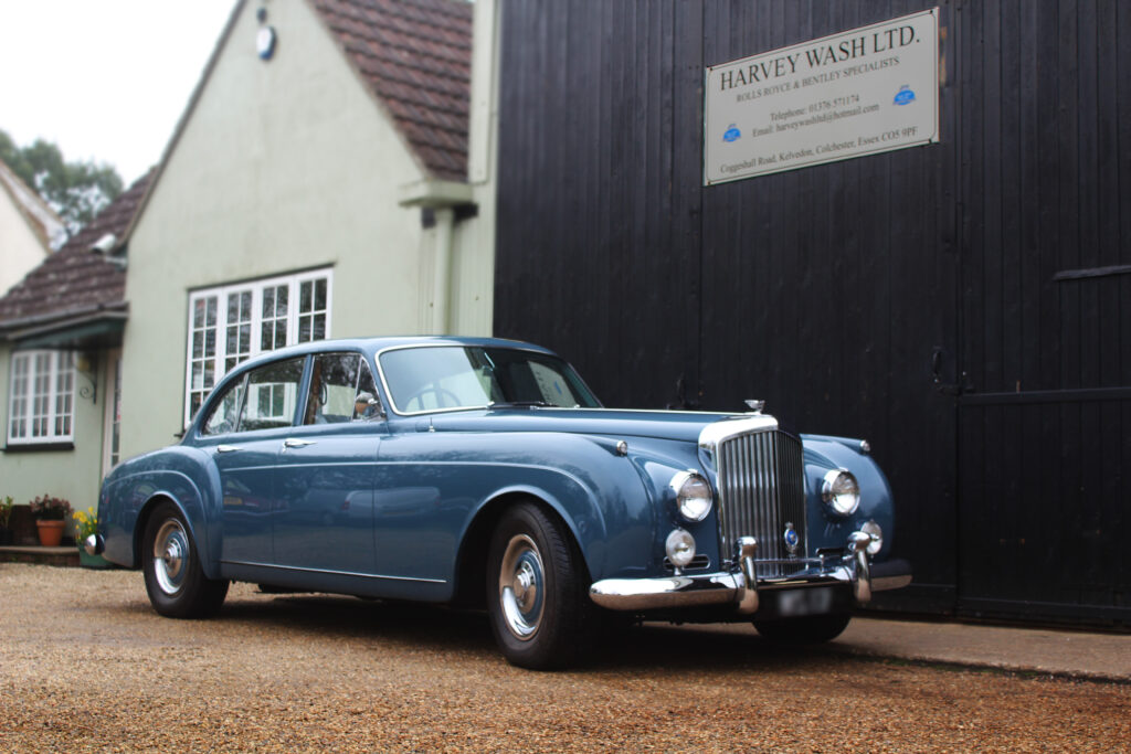 Pastel blue classic Bentley S1 continental, parked on a beige gravel driveway in front of a black door and pastel green building