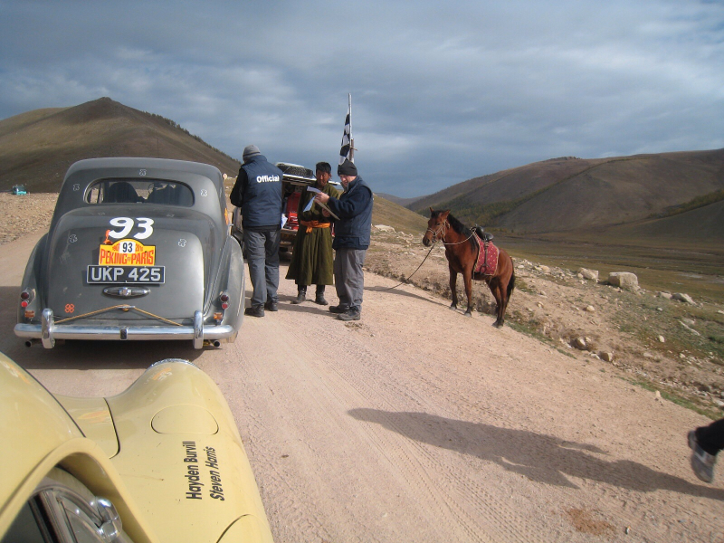 Rolls Royce Silver Dawn viewed from rear on Left hand side of road, parked at a checkpoint on a mountain road on the Peking to Paris rally. To the right hand side is a groupf of checkpoint officials and a brown horse.