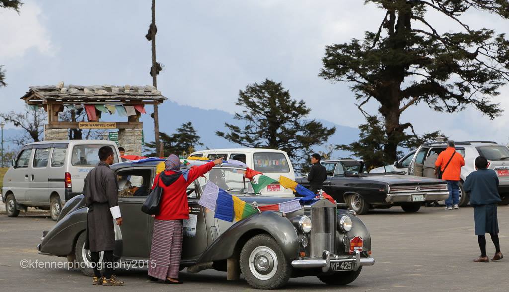 Rolls royce silver dawn rally car covered in coloured flags, in the background is a burmese building and someone has stopped to give directions 